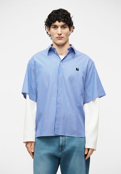 Ung mann med krøllete hår iført en kortermet skjorte med blå og hvite vertikale striper, over en langermet hvit skjorte og blå jeans.