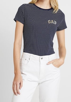 Kvinde iført en marineblå og hvid stribet kortærmet t-shirt med "GAP" logo og hvide bukser med høj talje, den ene hånd i lommen.