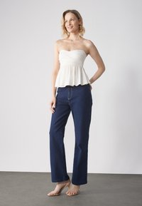 Top bianco senza spalline con corpetto arricciato, abbinato a jeans a gamba larga blu navy. La modella indossa sandali con tacco nudo.
