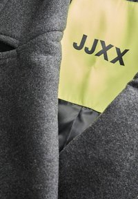 Sivi pamučni sako s mekom teksturom. Unutrašnjost ima svijetlu žutu podstavu s "JJXX" u podebljanim crnim slovima.