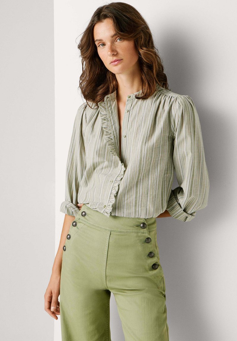 Junge Frau mit welligen braunen Haaren, die eine grün gestreifte Bluse mit Rüschen-Details und hoch taillierte hellgrüne Hose mit Knöpfen trägt.