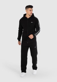 Felpa nera con zip e righe laterali bianche, abbinata a pantaloni neri coordinati. Presenta una tasca anteriore e sneakers bianche.