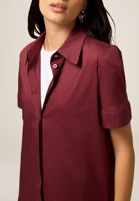 Bordeaux Kurzarmhemd aus glattem Stoff, mit klassischem Kragen, Knopfverschluss und aufgerollten Ärmeln.