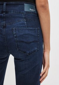 Sötétkék farmernadrág bőr logópárnával a derékrészen és egy hátsó zsebben ívelt varrásmintával.