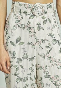 Bloemenprint broek van lichtgewicht stof, met een brede riem met een ronde gesp, patronen van groene bladeren en roze bloemen op een gestreepte achtergrond.