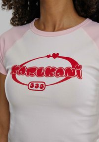White T-shirt with pink raglan sleeves, featuring red text "KARL KANI 333" and heart accents on the chest area. Soft cotton material.