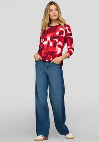 Frau trägt rotes, abstrakt gemustertes Langarmshirt, weit geschnittene blaue Jeans und weiße Sneaker, steht mit den Händen in den Taschen vor grauem Hintergrund.