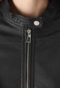 Giubbotto in pelle nera con colletto alto, dotato di zip argentata e chiusura con bottone a pressione. Superficie texturizzata con dettagli di design minimalisti.