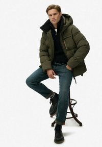 Groene gewatteerde jas met capuchon over een zwarte trui, gecombineerd met blauwe spijkerbroek en zwarte veterlaarzen. Zichtbare ritssluiting en logodetail.