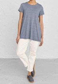 Grå t-shirt med kort ärm, lös passform och rundad halsringning med knappar på sidan, som är i kombination med krämfärgade byxor och gråa slipskor.