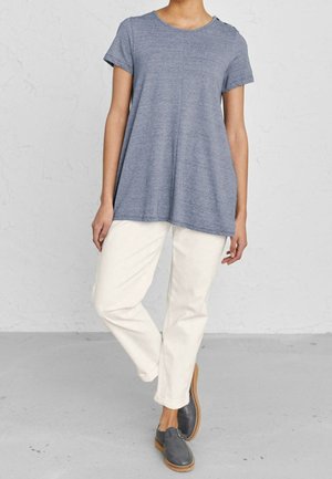 Camiseta gris de manga corta, holgada, con cuello redondo y detalles de botones en los laterales, combinada con pantalones crema y zapatos de tela gris sin cordones.