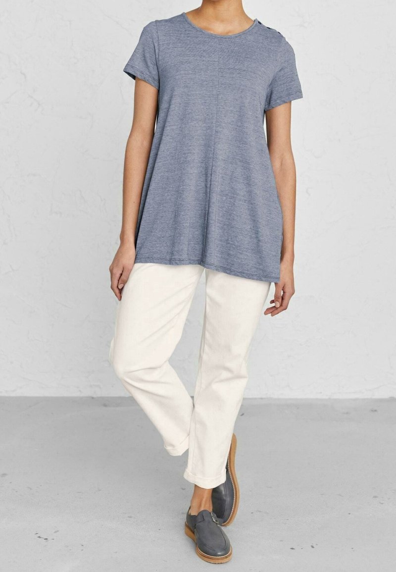 Grå t-shirt med kort ärm, lös passform och rundad halsringning med knappar på sidan, som är i kombination med krämfärgade byxor och gråa slipskor.