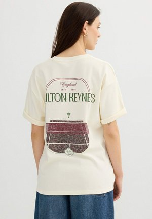Mujer con cabello largo y castaño, vistiendo una camiseta crema con mangas remangadas y un gráfico de la pista de tenis "Milton Keynes" en la parte trasera.
