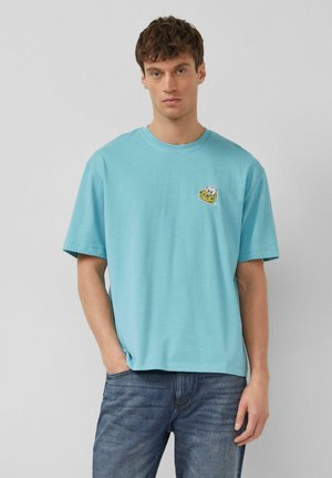 Młody mężczyzna w jasnoniebieskiej koszulce z małym żółtym logo tygrysa i niebieskich dżinsach, stojący na tle jednolitego szarego tła.