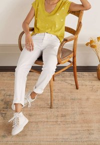 Žlutá košile s krátkým rukávem a vzorem listů; bílé, úzké džíny; bílé tenisky se zlatými detaily; sedící na dřevěné židli.