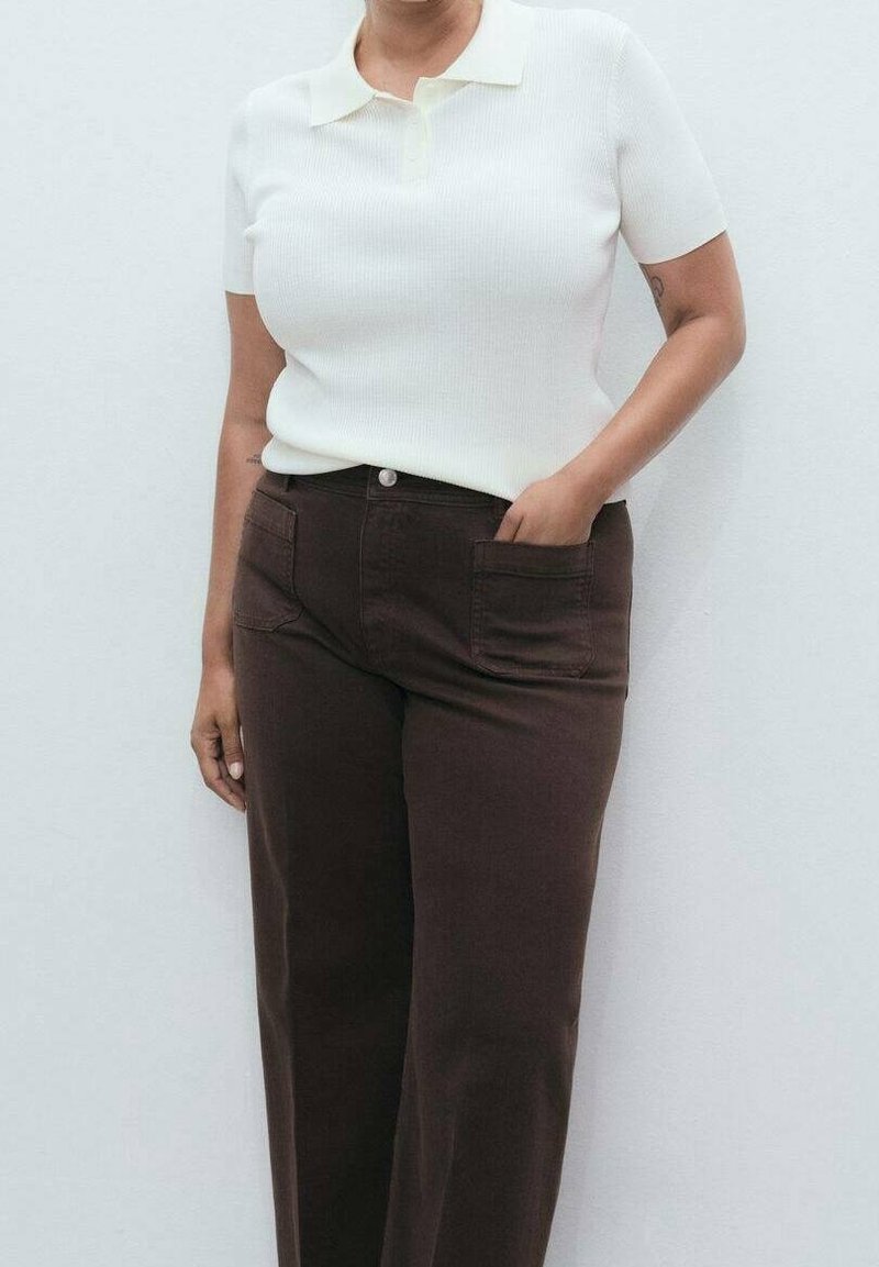 Personne portant un polo blanc côtelé à manches courtes rentré dans un pantalon marron foncé taille haute, les mains dans les poches, debout contre un mur blanc.