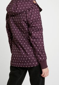 Lila jacka tillverkad av smidigt material, med ett geometriskt mönster som har små ljusrosa former och elastiska muddar.