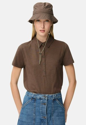 Femme portant un polo marron à boutons, un chapeau seau marron assorti avec cordon sous le menton, et une jupe en denim bleu à taille haute avec boutons sur le devant.