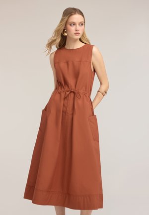 Jeune femme aux longs cheveux blonds portant une robe midi sans manches couleur rouille avec poches et ceinture nouée, regardant sur le côté sur un fond uni.