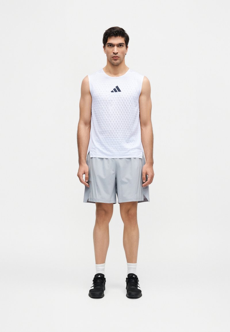 Homme debout face à l'avant, portant un débardeur blanc Adidas, un short gris clair, des chaussettes blanches et des chaussures de sport noires.