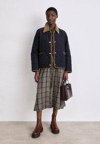 Casaco matelassado azul-marinho com colarinho de corduroy na cor areia, combinado com um vestido midi xadrez, botas de cano curto castanhas e uma pequena bolsa castanha escura.