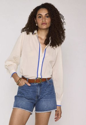 Donna con capelli ricci che indossa una camicetta crema con bordi blu, pantaloncini di jeans con orli sfilacciati e una cintura marrone, in piedi con una mano in tasca.