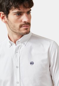Camisa blanca de botones de algodón con cuello estructurado, que presenta un pequeño logo azul bordado en el lado izquierdo del pecho. Textura suave.