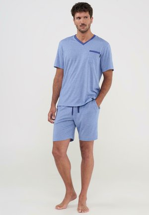 Hombre descalzo de pie, vistiendo una camisa de manga corta azul claro con cuello en V y bolsillo, y pantalones cortos a juego con cordón, frente a un fondo liso.