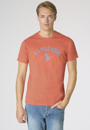 Giovane uomo con capelli biondi che indossa una t-shirt corallo U.S. Polo Assn. e jeans blu, in piedi con una mano in tasca su sfondo bianco.