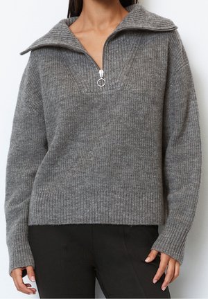 Kvinde iført en grå ribstrikket sweater med halv lynlås og bred krave, kombineret med sorte bukser, stående mod en hvid baggrund.