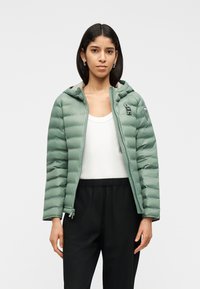 Γυναίκα με ίσια σκουρόχρωμη τρίχα φοράει ανοιχτό παλτό φουσκωτό ανοιχτού πρασινού χρώματος, λευκή φανέλα χωρίς μανίκια, μαύρο παντελόνι και ασημένια σκουλαρίκια κρίκους σε λευκό φόντο.