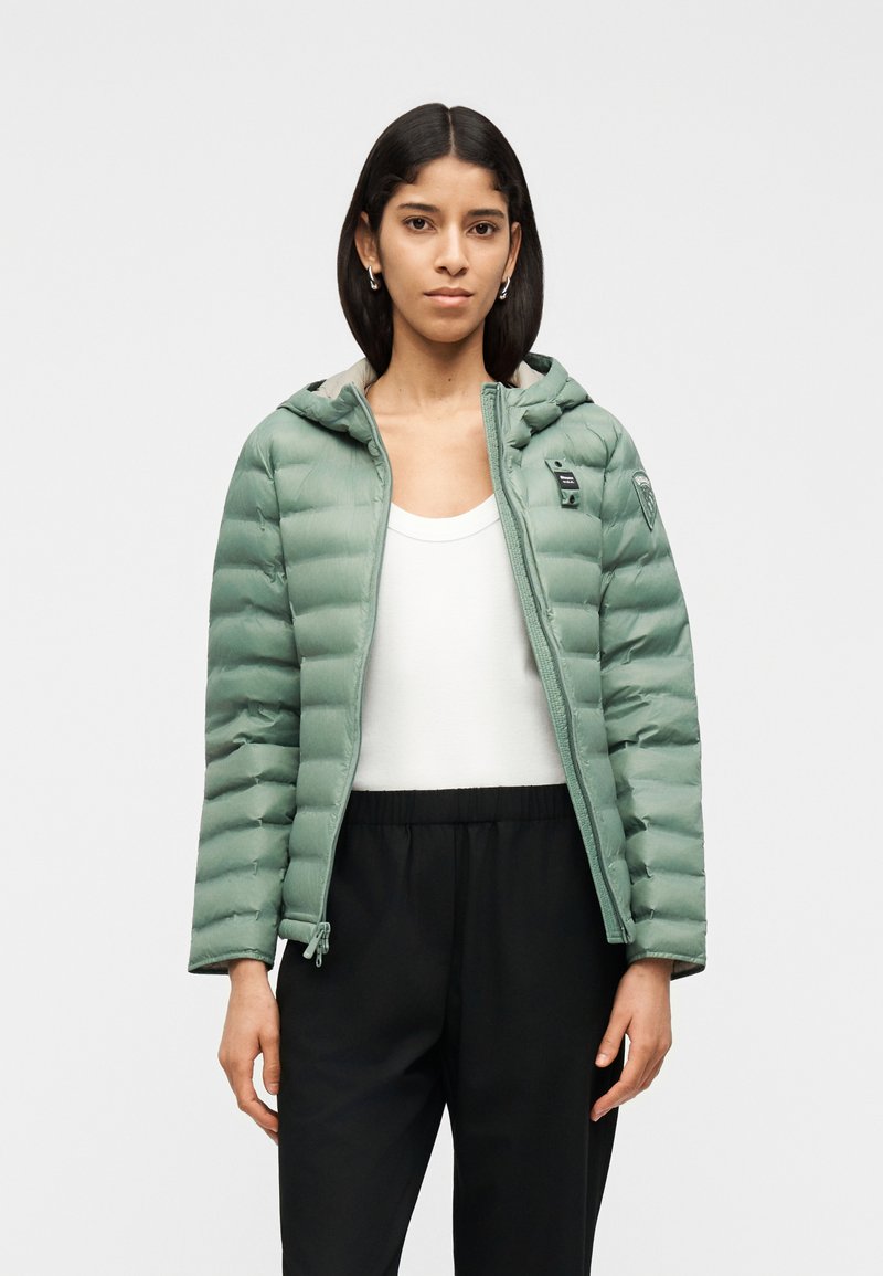 Γυναίκα με ίσια σκουρόχρωμη τρίχα φοράει ανοιχτό παλτό φουσκωτό ανοιχτού πρασινού χρώματος, λευκή φανέλα χωρίς μανίκια, μαύρο παντελόνι και ασημένια σκουλαρίκια κρίκους σε λευκό φόντο.