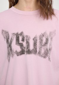 Rosa bomull t-skjorte med et stort, slitt svart grafisk trykk av ordet "KSUBI" på brystet. Rund hals, avslappet passform.