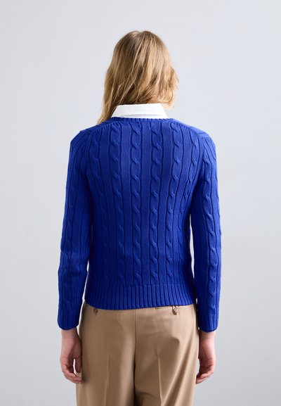 Zils adītais džemperis ar ribotu apakšmala un garām piedurknēm, uzvilkts virs baltas krekla ar apkakli; plakanas adīšanas tekstūra un pieguļošs dizains.