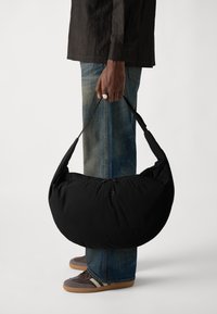 Borsa a spalla nera imbottita a forma di mezza luna, portata da una persona. Presenta una chiusura con zip e un tessuto resistente. Indossata con jeans casual e sneaker.