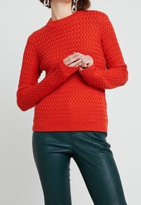 Pull en tricot rouge avec un motif texturé, col rond et poignets côtelés, associé à un pantalon en cuir vert.