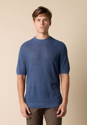Jonge man staat, gekleed in een blauw gebreid T-shirt met korte mouwen en een bruine broek, tegen een effen beige achtergrond.