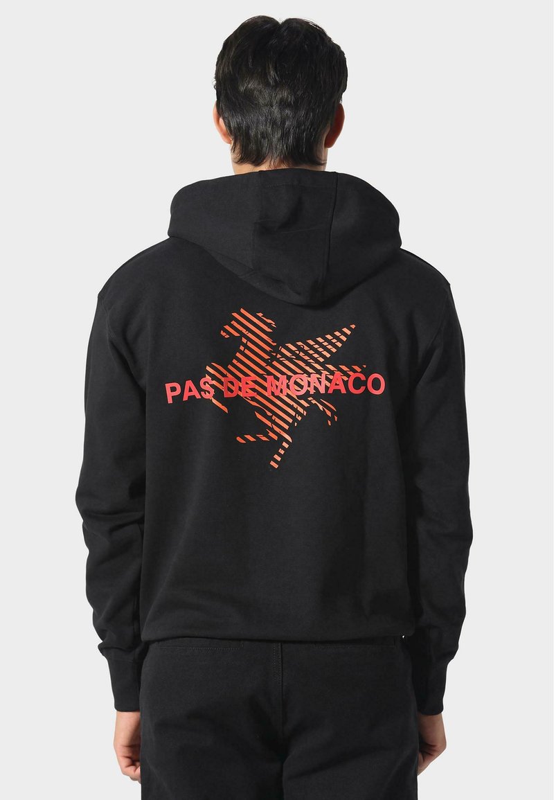 Sudadera con capucha negra que presenta un gráfico a rayas naranjas de un caballo y el texto "PAS DE MONACO" en la parte posterior. Tejido suave, corte relajado.