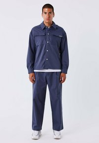 Marineblauwe jas met twee borstzakken en knoopsluiting, gecombineerd met bijpassende broek. Gladde stof, relaxte pasvorm en witte sneakers.