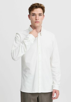 White long-sleeve button-up shirt with a collar, featuring a textured fabric and a straight hem, worn with a neutral-colored trouser.