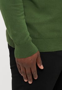 Zelený pletený sveter s rebrovanými manžetami, mierne priliehavý strih a hladká textúra. Čierne nohavice sú čiastočne viditeľné v popredí.