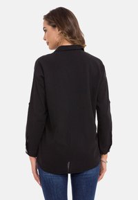 Zwarte langemouwen shirt met een ontspannen pasvorm, voorzien van een achterpand, met knoopmanchetten en een licht gebogen zoom. Gladde textuur.