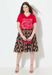 Rotes V-Ausschnitt-T-Shirt mit weißem Aufdruck, kombiniert mit einem schwarzen Rock mit orangenen Blumenmustern und grünem Saum. Hält eine rote Clutch.