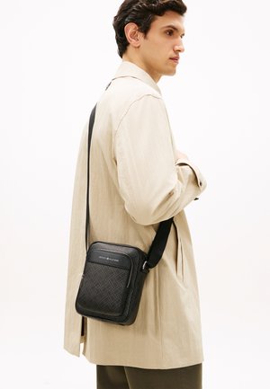 Mann iført beige jakke og olivengrønne bukser bærer en svart Tommy Hilfiger skulderveske med mønstrete overflate og frontlomme med glidelås.