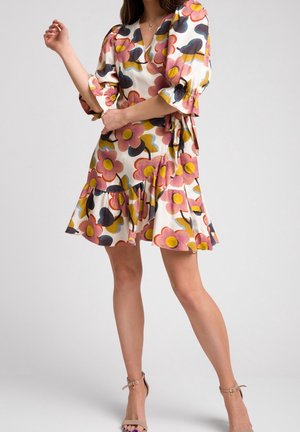 Kobieta ubrana w kwiecistą sukienkę kopertową z bufkowymi rękawami i falbaną u dołu, w połączeniu z beżowymi szpilkami na kostce na jednolitym tle.