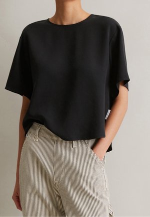 Person wearing a loose black short-sleeve top paired with beige and black vertical striped pants, one hand in pocket against a plain background.