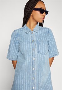 Lys blå denimkjole med vertikale hvite striper, korte ermer, krage og en brystlomme. Har sølvfargede knapper.