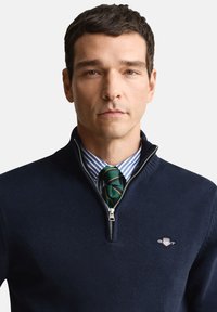 Marineblauwe trui met ritsnek, gemaakt van textuurgebreide stof, voorzien van een zilveren rits en een klein logo. Draagt over een blauw-wit gestreept overhemd en een groene stropdas.