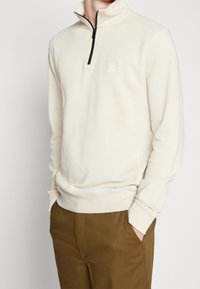 Světle béžová mikina na zip s vysokým límcem, která má decentní nášivku loga a žebrované manžety, kombinovaná s olivovými kalhotami.