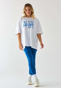 Biała, oversize'owa koszulka z niebieskim napisem i gwiazdkami, w zestawie z niebieskimi legginsami i białymi sneakersami z teksturowaną podeszwą oraz prążkowanymi skarpetkami.